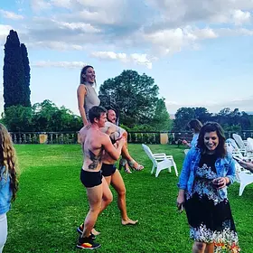 Shirtless male entertainer carrying a laughing guest on his shoulders during a fun outdoor party on a large grassy lawn.