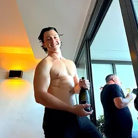 Drinks Duty With A Smile Smiling topless waiter in black pants holding a bottle of sparkling wine while helping host a relaxed private party.
