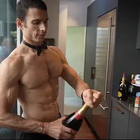 Topless waiter wearing a black bow tie opening a bottle of sparkling wine in a modern kitchen ready to serve party guests.