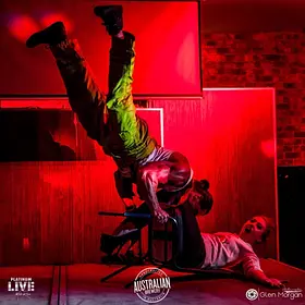 Male stripper in a firefighter costume performing an upside-down acrobatic stunt over a seated guest during a high-energy live stage show.