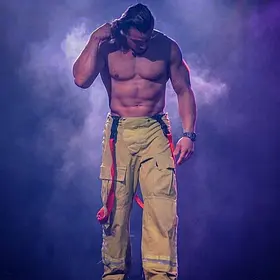 Firefighter Reveal Shirtless male stripper standing under smoky stage lighting in yellow firefighter pants as he prepares for a live performance.