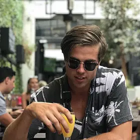 Tyler’s Sunny Side Sunshine Coast entertainer Tyler wearing sunglasses and a patterned shirt while enjoying an orange juice at a relaxed outdoor café.