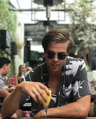 Sunshine Coast entertainer Tyler wearing sunglasses and a patterned shirt while enjoying an orange juice at a relaxed outdoor café.