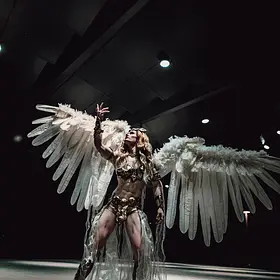 Queensland drag queen Tara Zaan commanding the stage in an elaborate fantasy costume with enormous white feathered wings spread beneath dramatic lighting.