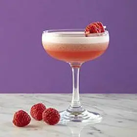 Pink raspberry drop martini with creamy foam served in a coupe glass on a marble surface with fresh raspberries and a purple background, styled for fruity cocktail recipe blogs, girls’ night drinks and cocktail making class inspiration.