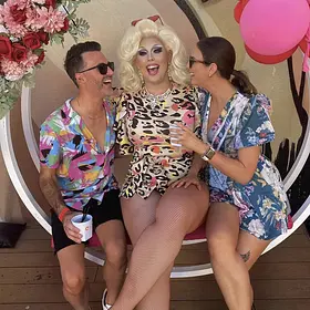 Alt Text: A blonde drag queen in a leopard-print dress laughing while sitting on a circular outdoor swing chair with two party guests at a daytime event.