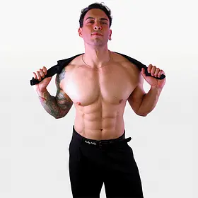 A muscular, shirtless male performer with a large tattoo on his right arm posing while holding a black shirt behind his neck against a white background.