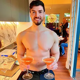 A smiling topless waiter with a beard holding two orange cocktails in salt-rimmed martini glasses in a modern kitchen setting.