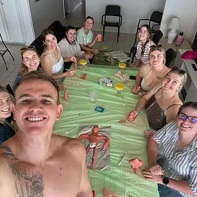 Hens Party Clay Workshop Group. A high-angle selfie of a muscular, shirtless male model smiling with a group of women around a table covered in green plastic, engaged in a creative clay sculpting workshop at a hens party.