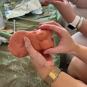 Clay Sculpting Close-Up. A close-up shot of hands covered in terracotta-coloured clay shaping a small sculpture of a female torso during a creative workshop.