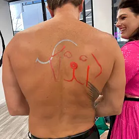A view from behind of a shirtless male host whose back has been painted with a simple red and silver outline of a dog, while a woman in a bright pink dress laughs nearby.