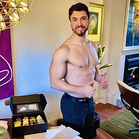 A smiling, muscular, shirtless male host giving two thumbs up while standing in a living room next to a box of colorful cupcakes.