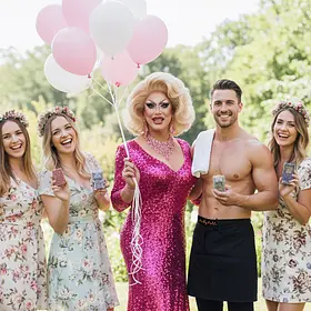 A drag queen in a pink sequinned dress holding balloons, standing outdoors with a shirtless male host and three women in floral dresses who are holding bars of soap.