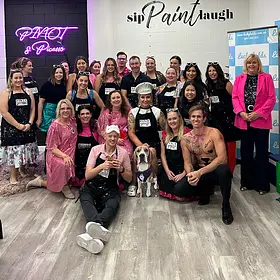 A large group of guests, many wearing pink and black aprons, pose with a shirtless male entertainer and a man in a pink polo shirt at a "Sip Paint Laugh" event held at a Pinot & Picasso studio.