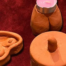 A collection of handmade terracotta clay sculptures including a heart-shaped tray, a round base with a phallic center, and a butt-shaped candle holder containing a pink tea light.