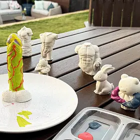 The "Buff Bear & Friends" A creative outdoor plaster and paint workshop setup featuring a bright lime green hand-painted phallic sculpture on a white plate alongside various unpainted naughty moulds, including muscular torsos, a rabbit, and a "buff" teddy bear. These cheeky craft sessions are available Australia-wide for hens parties and birthdays.