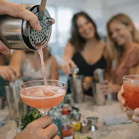 A close-up action shot of a cocktail being poured from a metal shaker into a coupe glass, with a group of women blurred in the background smiling and holding drinks during a cocktail making class. The image captures the hands-on experience, fresh cocktails, and social atmosphere of a hens party activity.