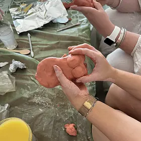 A close-up view of two hands shaping a soft piece of clay into a playful, risqué sculpture on a table covered with a green protective sheet. The person’s hands are lightly dusted with clay, showing the texture and messiness of the process as they press and smooth the surface. Around the workspace are small sculpting tools, bits of unused clay, a glass of a light-coloured drink, and crumpled foil, suggesting an informal, creative setting. Another person’s hands can be seen in the background working on their own piece. The scene captures a fun, hands-on hens party activity where guests laugh and get creative while sculpting cheeky clay designs together.