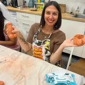 A smiling woman sits at a workshop table covered in a clear plastic sheet, holding up two small clay creations in her hands as if proudly showing them off. She wears a patterned apron with bold colourful text and a brown top underneath. Her arms are slightly raised, palms open, presenting each piece—one shaped like a small rounded sculpture and the other like a short cup with uneven, hand-formed edges. The table in front of her is messy with smears of orange clay, and a pack of wipes sits nearby. In the background, a clean kitchen-style workspace is visible with white cabinets, a wooden countertop, a sink, and small containers. Another person sits partially out of frame on the left, also working with clay. The overall feeling is relaxed and playful, capturing a moment of pride and laughter during a hands-on creative class.