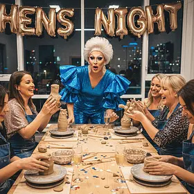 Hens Night Clay Workshop with Drag Host – Lucky Ladds A lively group of women sit around a wooden table shaping clay while laughing and chatting, each working on a small sculpted piece placed on a round board in front of them. In the centre, a drag host wearing a bright blue outfit with dramatic puffed sleeves and styled white hair stands leaning forward, reacting animatedly to the group’s creations. Several participants hold up their clay pieces to show each other, smiling and laughing. The table is scattered with clay tools, small chunks of clay, bowls, and glasses of sparkling drinks, along with bits of colourful confetti. Behind them, large windows reveal a nighttime city skyline, and gold balloon letters spelling “HENS NIGHT” hang above. The scene captures a high-energy, social workshop atmosphere filled with humour, creativity, and celebration.