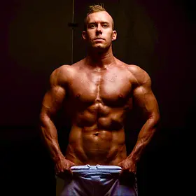 A muscular adult man stands facing forward in a dark, studio-like setting with warm, low lighting that highlights the contours of his chest, shoulders, and abdominal muscles. He is shirtless and holds the waistband of his light-coloured pants with both hands, pulling them slightly forward. His expression is serious and focused as he looks directly at the camera. The background is deep and shadowed, creating a high-contrast, dramatic effect that draws attention to his physique and posture.