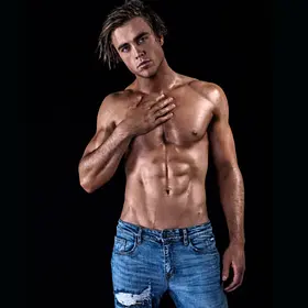 A shirtless adult man with a muscular, athletic build stands against a plain black background. He wears light blue denim jeans with a slightly distressed patch on one thigh. His body is angled slightly toward the camera, with one hand resting flat against the upper left side of his chest and the other arm relaxed by his side. His hair is styled loosely and swept to one side, and he looks directly at the camera with a calm, confident expression. Soft lighting highlights the definition in his chest, shoulders, and abdominal muscles, while the dark backdrop keeps the focus entirely on his pose and physique.