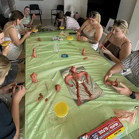 At a relaxed indoor hens party, a group of women sit around a long table covered with a green plastic tablecloth, shaping clay into playful sculptures. Each participant focuses on their own piece, using sculpting tools and their hands to form details. In the center of the table, a larger clay figure lies on foil as a reference or group creation. Drinks, tools, and small clay pieces are scattered across the table, creating a casual, hands-on workshop atmosphere. The scene captures a fun and creative group activity with participants chatting and laughing while they work.