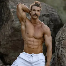 Topless Waiter Outdoor Rocky Pose – Lucky Ladds A shirtless male entertainer poses outdoors among large rocks, wearing light shorts and resting one arm behind his head. He has a muscular, athletic build and a styled moustache, creating a rugged yet polished promotional look in a natural setting.