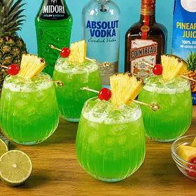 Fresh Midori Illusion cocktails ready to serve A vibrant cocktail styling scene featuring four Midori Illusion cocktails served over ice in rounded glasses with pineapple wedge and cherry garnishes.