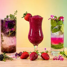 Three cocktails displayed side by side on a wooden surface, including a Blackberry Thyme Fizz with fresh blackberries and herbs in a tall glass, a Frozen Strawberry Daiquiri in a curved glass garnished with a strawberry, and a Sakura Mojito with mint, lime, and floral garnish in a highball glass. Fresh fruit, herbs, and small flowers are arranged around the drinks, with a soft pastel gradient background creating a vibrant tropical cocktail making class setting.