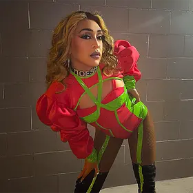Moesha Neon Pink And Green Performance Look Perth drag queen Moesha posing in a neon pink and green performance outfit with dramatic sleeves and thigh-high boots.