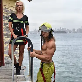 Firefighter-themed male stripper posing shirtless on a waterfront pier beside a female firefighter character, wearing firefighter pants, helmet and suspenders while showcasing a muscular physique.