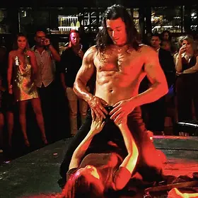 Male stripper performing an energetic interactive dance routine with a guest while a crowd watches from around the stage.