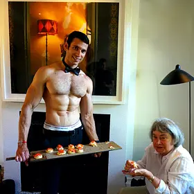 Smiling topless waiter wearing a black bow tie offering a tray of bite-sized food to a seated guest at a house party.