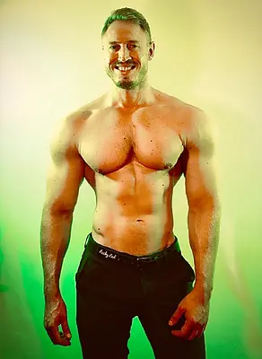 Smiling muscular shirtless man wearing black pants, posing against a softly lit yellow and green studio background.