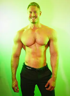 Adam smiling at the camera while posing shirtless in black pants against a bright green and yellow studio background.