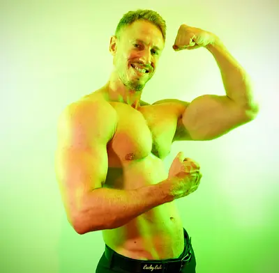 Adam smiling and striking a playful double-arm muscle pose shirtless against a vibrant green studio background.