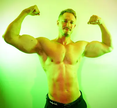 Adam grinning at the camera while flexing both arms shirtless against a bright green studio background.