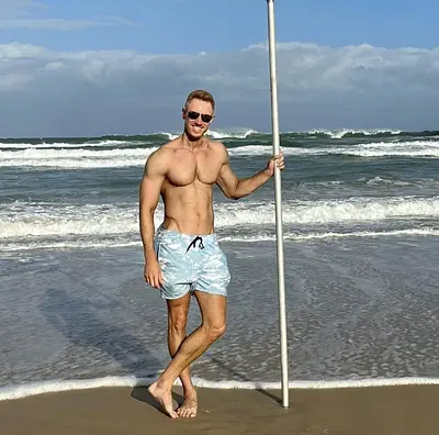 Shirtless male entertainer wearing sunglasses and light blue shorts standing beside a pole on a sunny beach with ocean waves behind him.