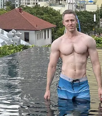 Shirtless male entertainer standing in an infinity pool wearing blue swim shorts, with resort buildings and tropical surroundings in the background.