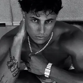 Liam posing shirtless in a black-and-white portrait with a pearl necklace, tattoos and wristwatch, available to book as a Canberra male entertainer.