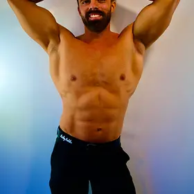 Gabriel smiling with both hands behind his head while posing shirtless in fitted black pants against a blue studio background.