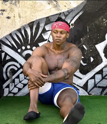 O’Niel sitting shirtless in blue athletic shorts and a red bandana against a bold mural, available to hire as a Sunshine Coast male entertainer.