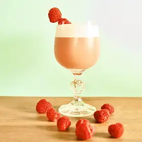 Creamy raspberry ice cream cocktail served in a vintage stemmed glass with frothy foam and fresh raspberry garnish, surrounded by scattered raspberries on a wooden table with soft pastel green lighting, perfect for dessert cocktail recipes, girls’ nights and cocktail making classes.