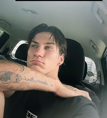 Ben sitting in a car wearing a black T-shirt, looking out the window with his tattooed arm resting across his chest.