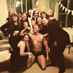Shirtless male entertainer kneeling in the centre of a laughing group of women during a lively hens party at home.